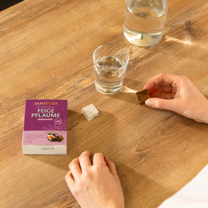 Person hält einen Fruit Cube Feige Pflaume in der Hand und sitzt an einem Tisch. Daneben steht ein Glas mit Wasser und die Fruit Cubes Verpackung